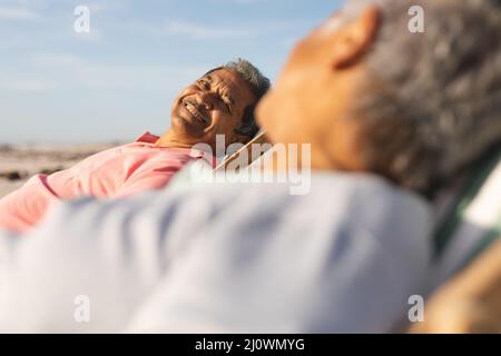 Lächelnder älterer Birazialmann, der auf einem Stuhl sitzt, während er die Frau anschaut, die sich am Strand entspannt Stockfoto