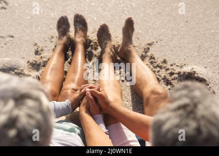 Blick aus dem hohen Winkel auf ein liebevolles, multirassisches Seniorenpaar, das die Hände am sonnigen Strand auf Sand hält Stockfoto