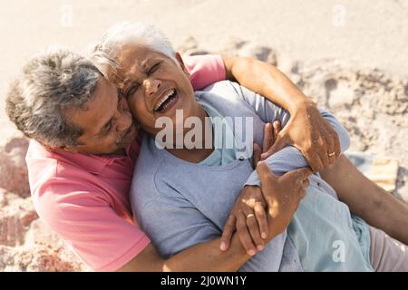 Der ältere Biracial-Mann umarmte eine fröhliche Frau von hinten, während er an einem sonnigen Tag am Strand saß Stockfoto