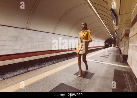 Auf der U-Bahn-Plattform spazieren schönes junges Mädchen mit langen Beinen in einem gelben Frühlingsmantel und einer weißen Handtasche in der Hand. Stockfoto