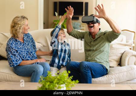 Glückliche kaukasische Großmutter und Enkel beobachten Großvater mit vr-Headset auf der Couch zu Hause Stockfoto