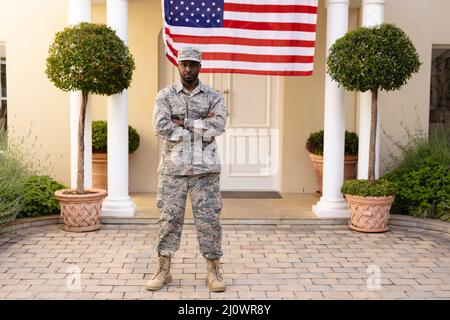 Porträt eines selbstbewussten afroamerikanischen Soldaten, der mit gekreuzten Armen auf dem Haus gegen die Flagge steht Stockfoto