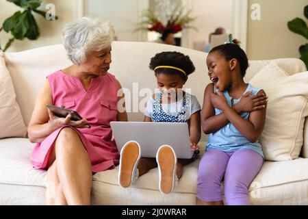 afroamerikanische Großmutter und ihre beiden Enkelinnen benutzen einen Laptop, der zu Hause auf der Couch sitzt Stockfoto