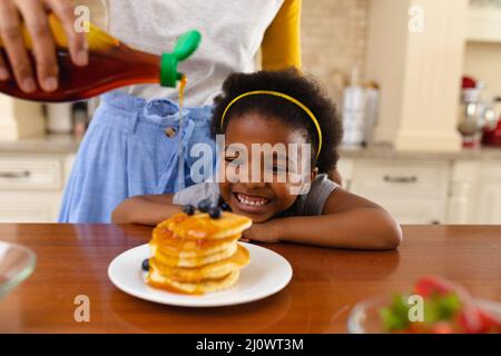 Mittengruppe der Mutter, die Ahornsirup auf den Pfannkuchen ihrer lächelnden afroamerikanischen Tochter zu Hause setzte Stockfoto