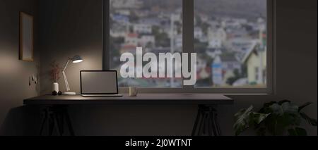 Moderner Home-Office-Arbeitsplatz am Abend mit Laptop-Computer White-Screen-Mockup, Licht von Tischlampe und Zubehör auf dem Tisch gegen den Windo Stockfoto