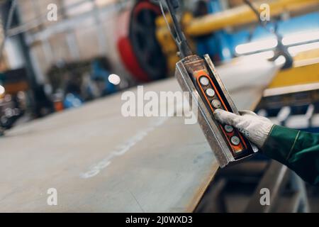 Arbeiter heben Metallblech mit Krankettenzug mit Fernbedienung und Haken in der industriellen Fabrik. Stockfoto