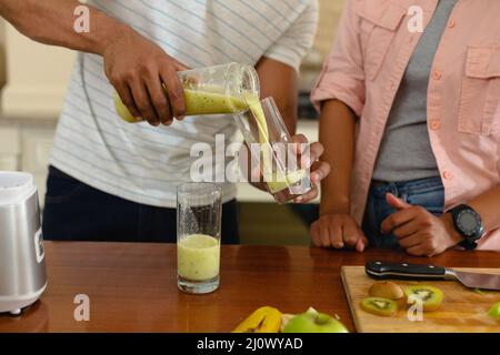 Mittelteil eines jungen afroamerikanischen Mannes, der auf der Kücheninsel ein Getränk aus dem Glas ausgießt Stockfoto