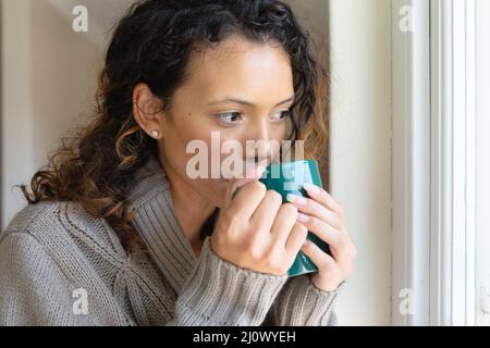 Junge Birazialfrau, die Kaffee aus dem Becher trinkt, während sie nach Hause schaut Stockfoto