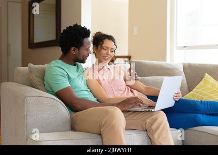 Lächelndes junges biracial Paar mit Laptop zusammen, während auf dem Sofa zu Hause sitzen Stockfoto