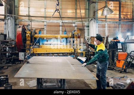 Arbeiter heben Metallblech mit Krankettenzug mit Fernbedienung und Haken in der industriellen Fabrik. Stockfoto