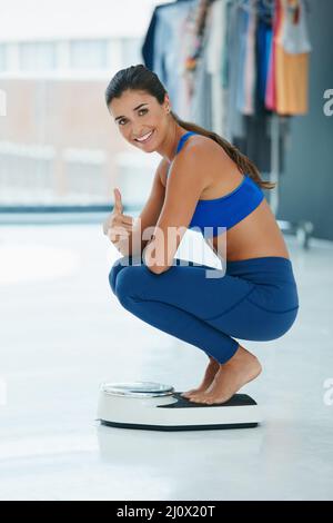 Meine Ziele zur Gewichtsabnahme sind auf dem richtigen Weg. Porträt einer fitgen jungen Frau, die sich auf einer Waage wiegt und den Daumen zeigt. Stockfoto