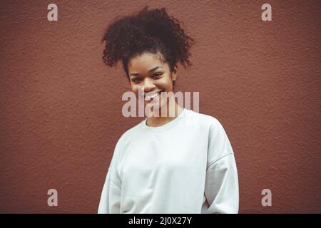 Reines Glück. Nettes glückliches afroamerikanisches Mädchen mit lockiger Frisur trägt weißen Pullover posiert gegen braune Wand Stockfoto