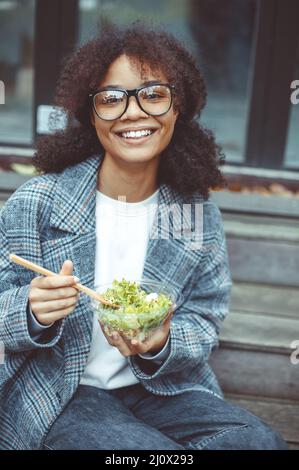 Glückliche afroamerikanische Büroangestellte, die draußen Salat isst, während sie auf der Bank im Park sitzt Stockfoto