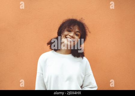 Reines Glück. Nettes glückliches afroamerikanisches Mädchen mit lockiger Frisur trägt weißen Pullover posiert gegen orange Wand Stockfoto