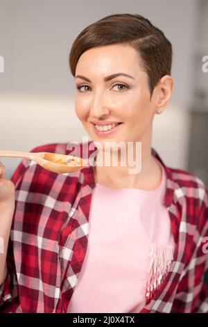 Nahaufnahme Hausfrau in roten karierten Hemd und kurze Haare Kochen Suppe in der modernen Küche. Lächelnde Frau testet ihr Gericht. Küchenchefin Stockfoto