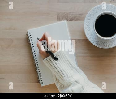 Frau, die in einem Notizbuch auf einem Holztisch schreibt, Hand in einem Pullover, der den Stift hält, Stockfoto