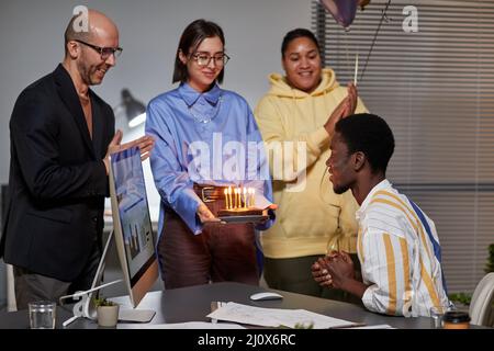 Verschiedene Gruppen von Menschen bringen Geburtstagskuchen mit Kerzen zu jungen Mann im Büro Stockfoto