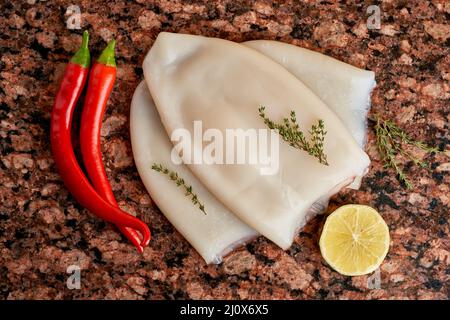 Drei Kadaver von frischem rohem Tintenfisch mit Zitrone, Pfeffer, Thymian auf einem dunklen Marmortisch. Stockfoto