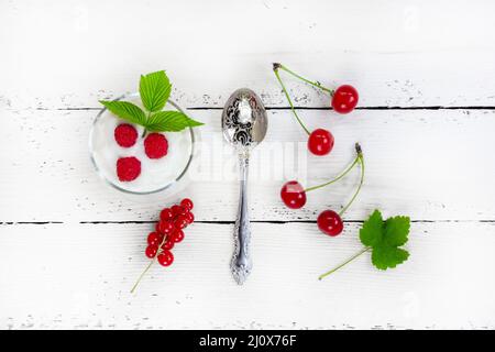 Ein Glas mit Milchjoghurt, reifen Himbeeren, einem grünen Blatt, roten Johannisbeeren und Kirschen auf einem weißen Holztisch mit einem Top-VI Stockfoto