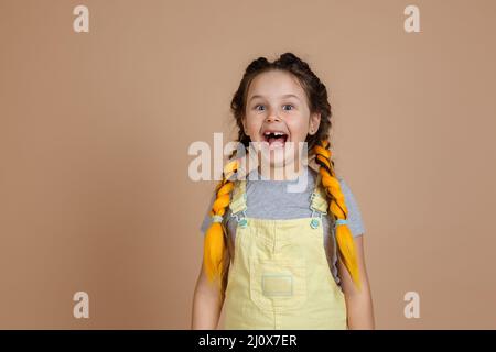 Verspielte, aufgeregte kleine Hündin mit gelben Kanekalon-Zöpfen, lächelnd mit geöffnetem Mund, mit fehlendem Zahn, gelbem Jumpsuit und grauem T-Shirt an Stockfoto