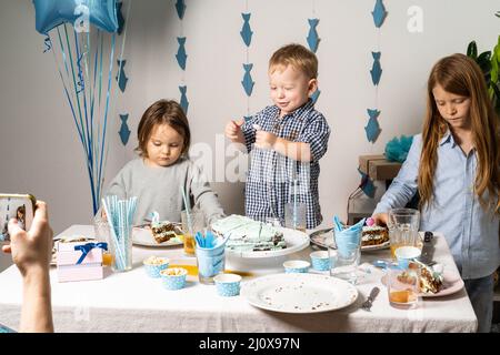 Brüder. Knabengeburtstag am Tisch. Die Jungs am Tisch schneiden einen Geburtstagskuchen. Mama fotografiert am Telefon den Geburtstag ihres Kindes. Stockfoto