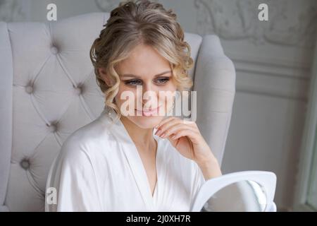 Hautpflege für Frauen. Kaukasische Frau berührt ihr Gesicht und sieht im Wohnzimmer in einem seidenweißen Bademantel in einem Sessel am Fenster im Spiegel aus. Genießt ihre Reflexion mit schönen Haaren und Make-up Stockfoto