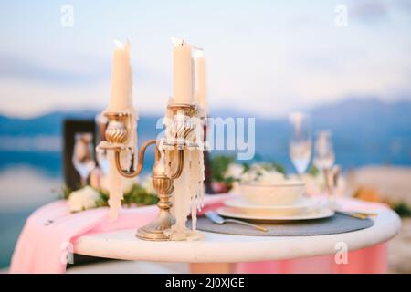 Nahaufnahme eines Hochzeitsessentischs an der Rezeption. Brennende Kerzen in einem Kerzenleuchter auf einem Tisch mit rosafarbenem Tuch und cremefarbenen Tellern Stockfoto