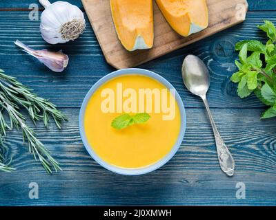 Cremige Suppe von Kürbis auf dunkelblauem Hintergrund Holz, Rosmarin, Knoblauch, Minze, kürbisscheiben. Ansicht von oben. Stockfoto