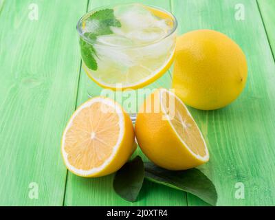 Limonade im Glas, eine Zitronenhälfte, frische Blätter auf dem grünen Tisch. Ein erfrischendes kaltes Getränk aus Wasser mit Eis, Minze und Scheiben o Stockfoto