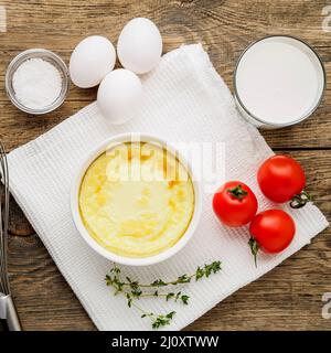 Weißes rundes Ramekin mit im Ofen gebackenem Omelett aus Eiern und Milch, mit Zutaten - Tomaten, Eiern, Salz, Glas Milch auf brauner r Stockfoto