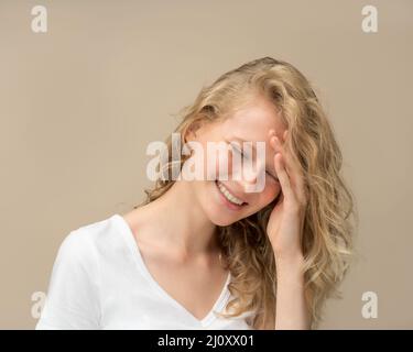 Schöne junge Mädchen lachen. Hübsche Blondine mit lockigen Haaren in weißem T-Shirt an beigefarbener Wand Stockfoto