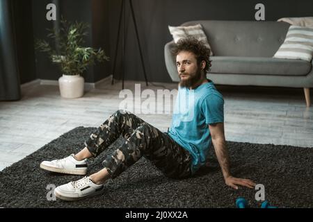 Hübscher junger Mann, der zuhause trainieren und entspannt auf dem Teppich sitzen und die Kamera betrachten kann. Lustige lockige Haare Mann mit einem Durcheinander Stockfoto
