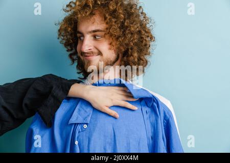 Junger Mann mit rothaarigen Locken lächelt, während er das Hemd isoliert über der blauen Wand probiert Stockfoto