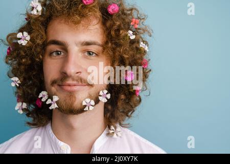 Nahaufnahme eines lächelnden jungen weißen Mannes im Hemd mit Blumen in seinem lockigen Haar und Bart, der auf blauem Hintergrund steht Stockfoto
