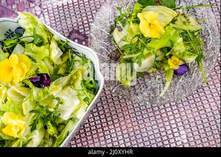 Frischer grüner Salat und Rucola-Salat mit Stiefmütterchen auf Glasplatte und in einer Schüssel auf violettem Hintergrund, Nahaufnahme. Stockfoto
