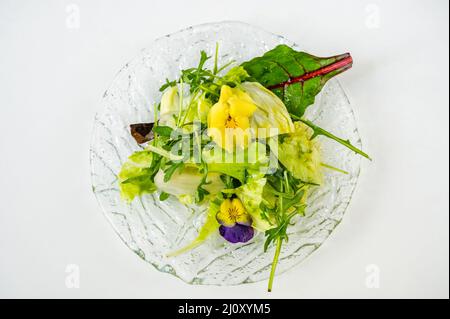 Frischer grüner Salat mit essbarem Stiefmütterchenkopf auf Glasplatte auf weißem Hintergrund, Nahaufnahme. Stockfoto