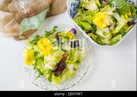 Frischer grüner Salat (Salat, Rucola, Roter Mangold) mit essbarer Stiefmütterchen-Blume auf Glasteller und in Schüssel, Schal auf weißem Hintergrund, Nahaufnahme. Stockfoto