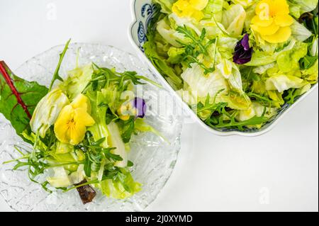 Frischer grüner Salat (Salat, Rucola, roter Mangold) mit essbarer Stiefmütterchen auf Glasplatte und in einer Schüssel auf weißem Hintergrund, Nahaufnahme. Stockfoto