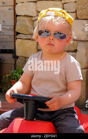 Molliger kleiner Junge, der draußen auf einem Spielzeugauto sitzt und eine Sonnenbrille mit einem leeren oder gelangweilten Ausdruck trägt Stockfoto