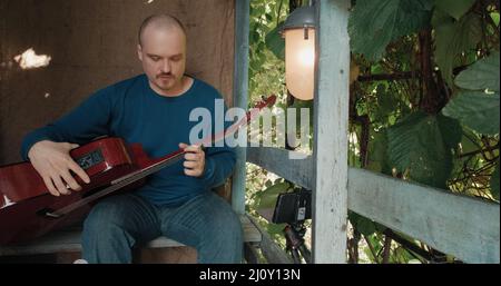 Der Mensch leitet eine Online-Akustikgitarre-Lektion von der Veranda seiner Vorstadtfarm aus Stockfoto