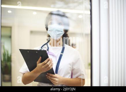 Junge müde überarbeiteten afroamerikanischen Arzt trägt eine schützende Gesichtsmaske mit Röntgen-Scan in den Händen Blick aus dem Fenster Stockfoto