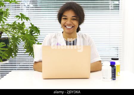 Junge glückliche afroamerikanische Ärztin, die am Arbeitsplatz sitzt und lächelnd auf die Kamera blickt, während sie auf einem Laptop in Modern arbeitet Stockfoto