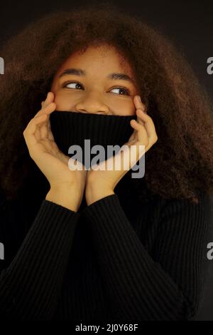 Erfreut afroamerikanische lockige Frau mit geschlossenen Augen bedeckt Gesicht mit warmen schwarzen Pullover Kragen Stockfoto