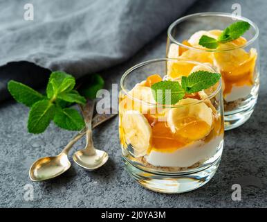 Gesundes mehrlagiges Dessert mit Joghurt, Banane, Mangomarmelade, Keks im Glas auf grauem Steingrund, Seitenansicht Stockfoto