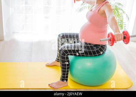 Junge Schwangere Hantelhanteln auf einem Fitnessball. Körperstraffung und Übungen während der Schwangerschaft Stockfoto