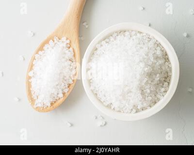 Weiße Schale mit großem Meersalz, Holzlöffel, verstreute Salzkristalle auf einem grauen Steintisch. Draufsicht, Nahaufnahme. Stockfoto