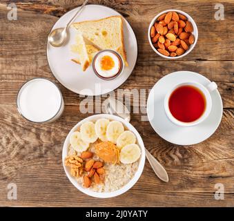 Frühstück am Morgen, auf einem Holztisch, Ansicht von oben. Ein Glas Milch, Kaffee, Müsli mit Früchten und Nüssen, weich gekochtes Ei. Stockfoto