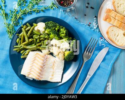 Gebackenes Kabeljaufilet mit Gemüse - Brokkoli, grüne Bohnen und Blumenkohl auf blauem Teller, Brot, blaue Serviette, Holzbac Stockfoto