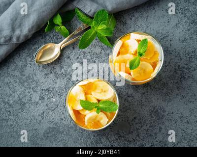 Gesundes mehrschichtiges Dessert mit Joghurt, Banane, Mangomarmelade, Keks im Glas auf grauem Steingrund, Draufsicht Stockfoto