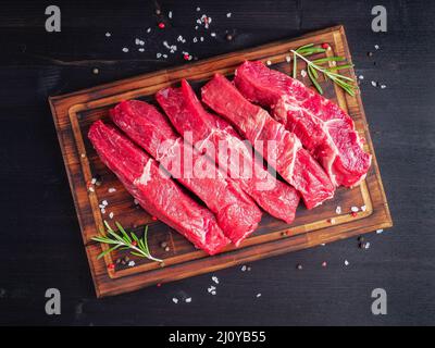 Rohes Fleisch, Rindersteak mit Würze auf Schneidebrett auf dunklem Hintergrund mit Rosmarin, Stockfoto